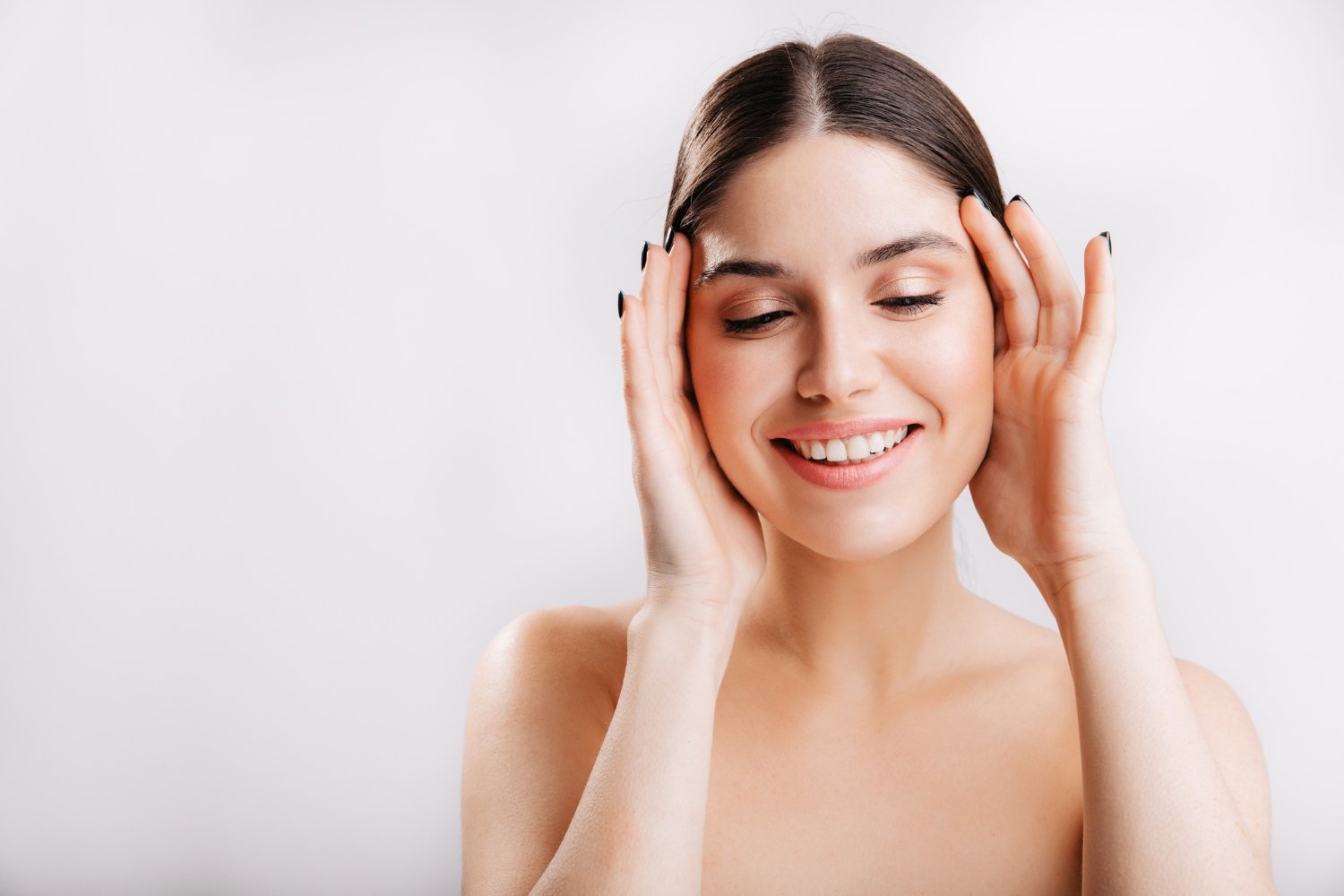 Mujer joven sonriente con la piel del rostro sin maquillaje, con las manos junto a las mejillas sobre fondo claro